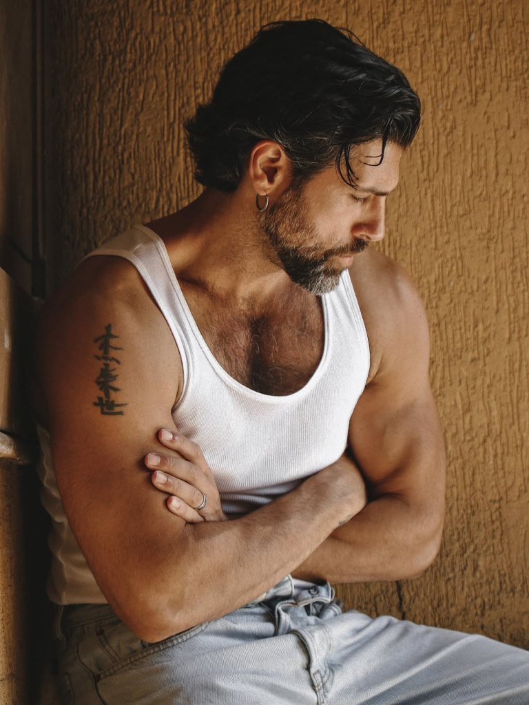 Italian actor Gilles Rocca poses in black-and-white and color portraits photographed by Marco Melfi in Testaccio, Rome, showcasing his hairy chest, muscular physique, and gray-bearded rugged masculinity.